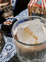 A glass of iced beverage with an ice cube featuring the "REVIVAL" logo sits on a table, with a blurred background showing what appears to be a puppet box and small boxes with harry potter poster on top